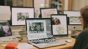 Uma mesa de trabalho com iMac, um Macbook, dois iPads e papéis espalhados pela mesa com o título "Pesquisa de Demanda por Soluções Digitais" e o subtítulo "Contribua para o Futuro do Marketing Digital"
